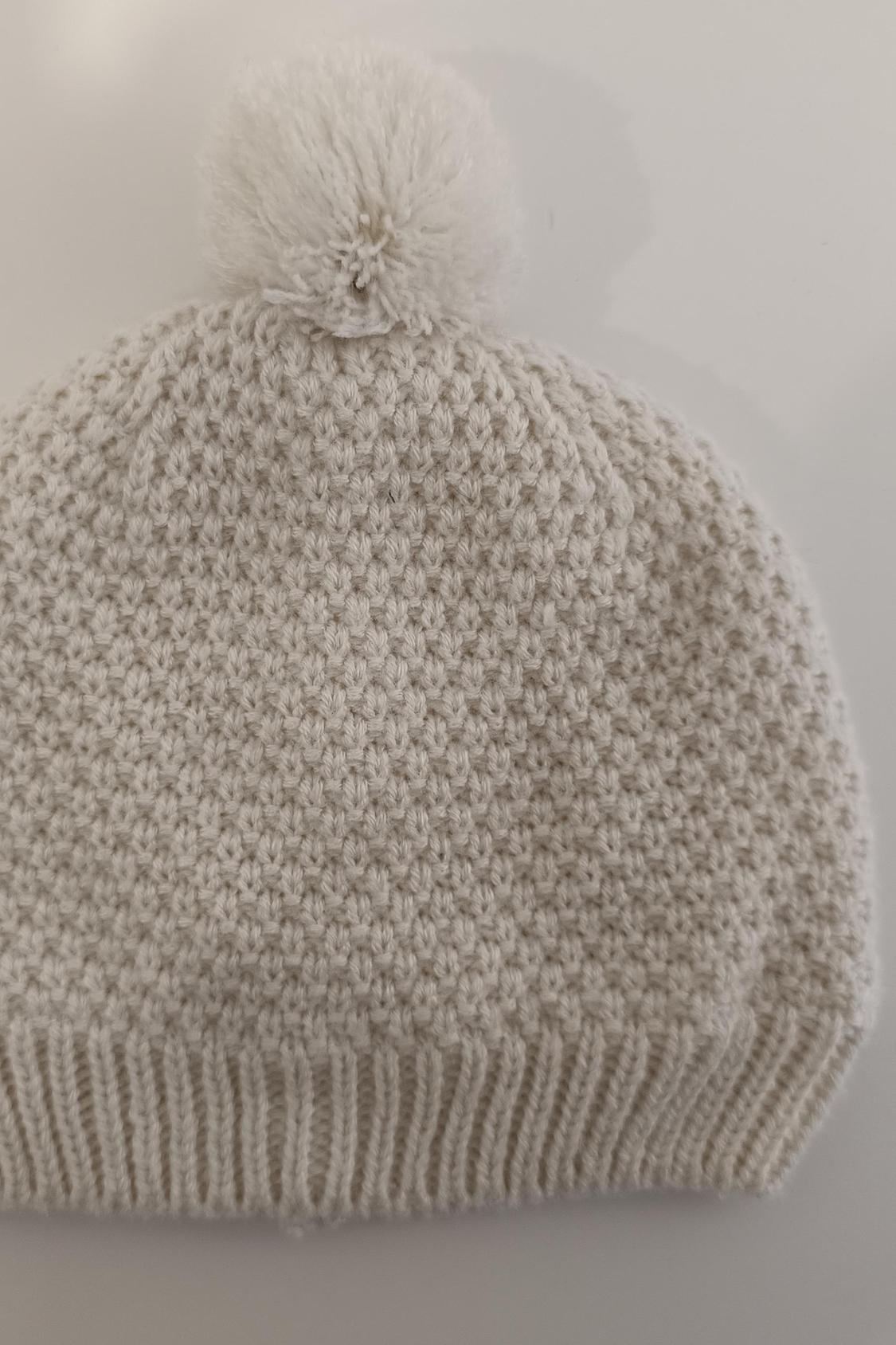 Detail of a cream-coloured knitted hat with a pom-pom on top.