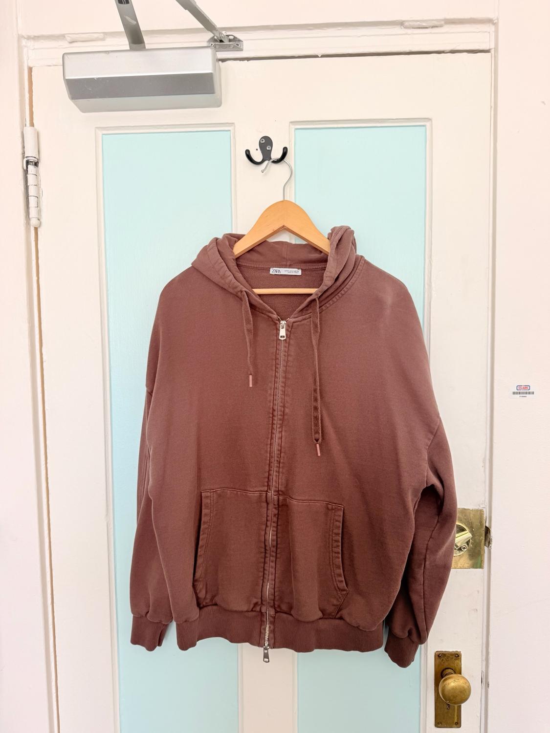 Brown hooded zip-up sweatshirt hanging on a white and light blue door. Text: ZARA. Casual and relaxed atmosphere.
