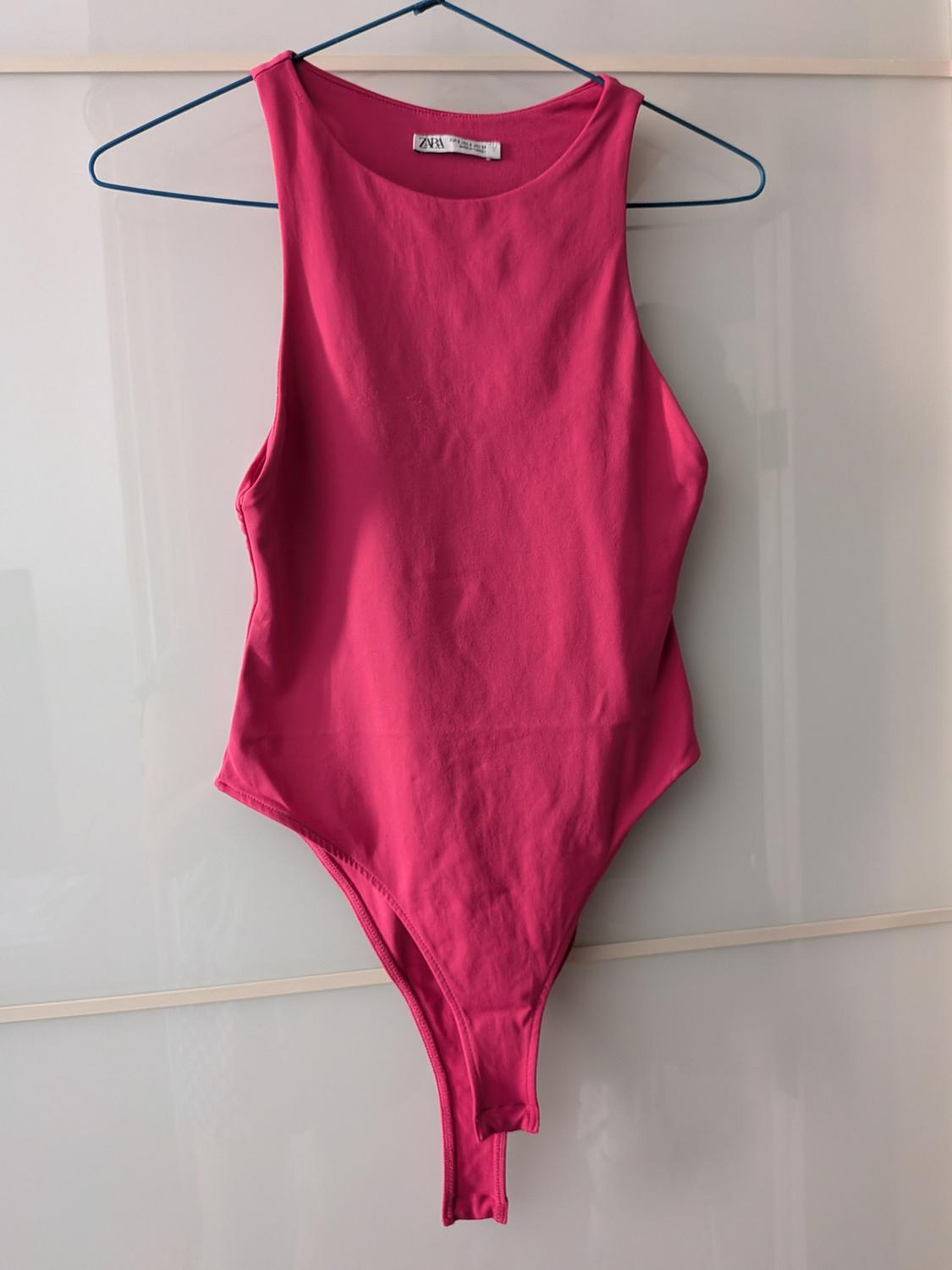 Fuchsia pink sleeveless high-neck bodysuit hanging on a blue hanger in front of a white wardrobe. Label with the text ZARA.