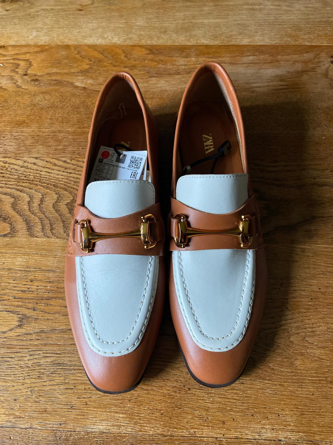 Brown and white loafers with gold metal detail on a wooden surface. Visible text: ZARA, EAC.