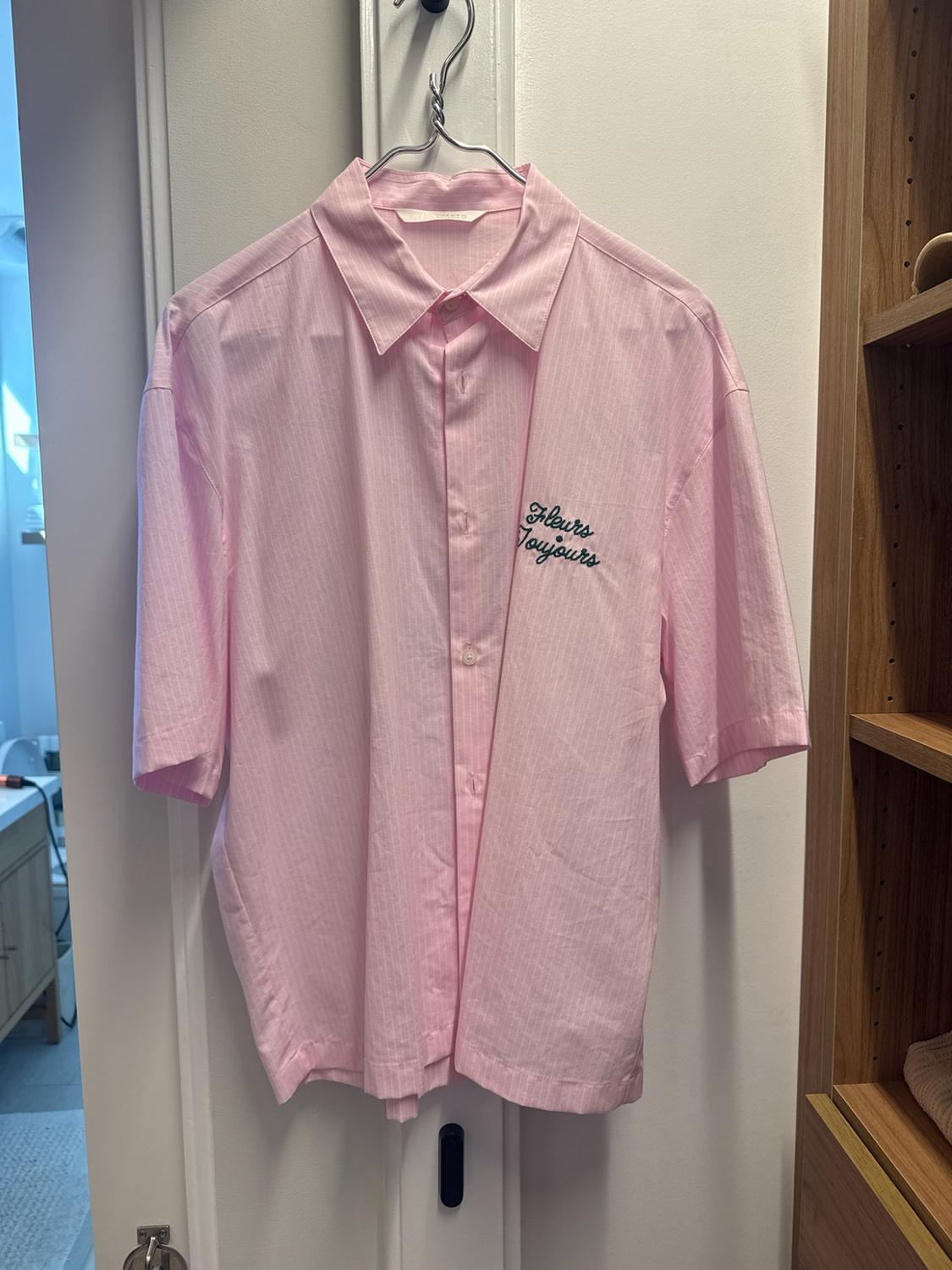 Short-sleeved pink shirt hanging on a door. On the chest it has the text "Fleurs Toujours". Relaxed home setting.