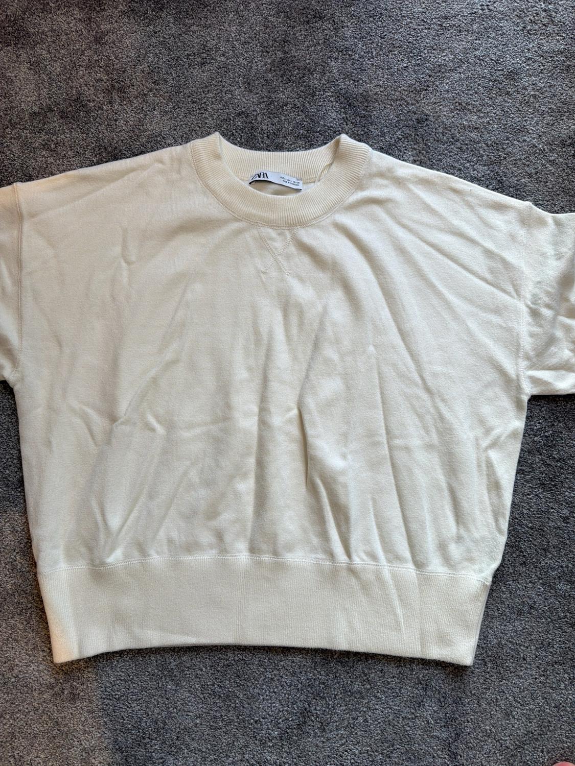 Cream short-sleeved sweatshirt with a ribbed crew neck and ZARA label, on a grey rug.