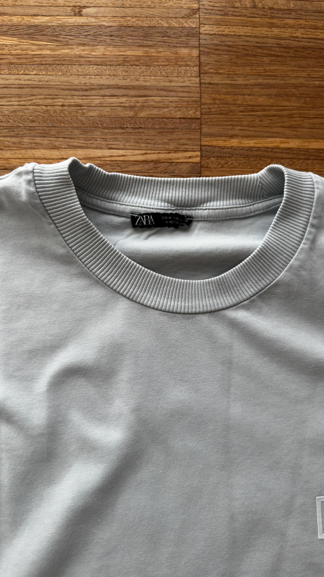 Κοντινό πλάνο ενός ανοιχτό γκρι t-shirt με ριμπ στρογγυλή λαιμόκοψη σε ξύλινο φόντο. Μαύρη εσωτερική ετικέτα με το κείμενο ZARA.
