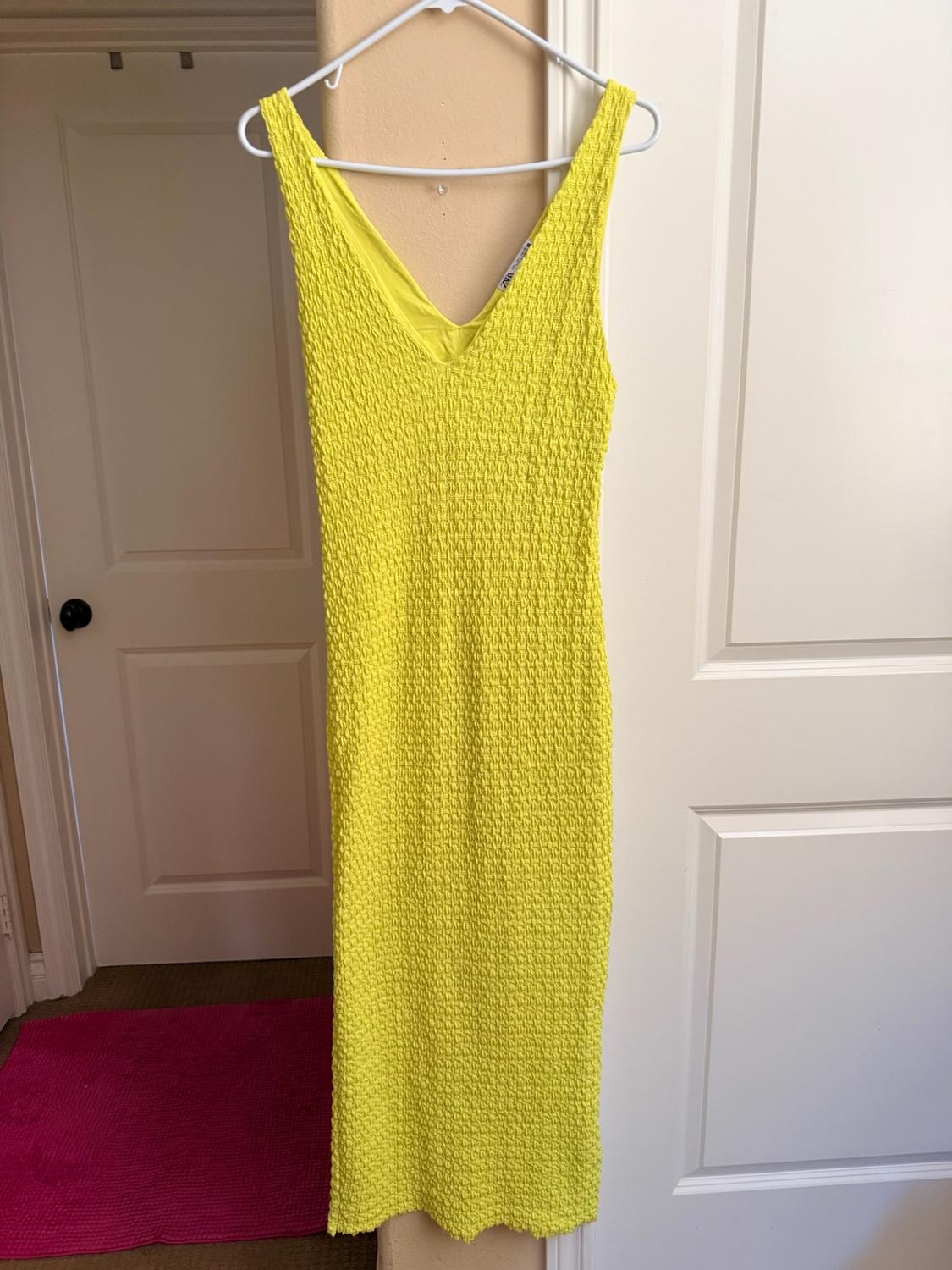 Textured yellow sleeveless dress hanging on a white hanger between two white doors. There is a pink rug on the floor.