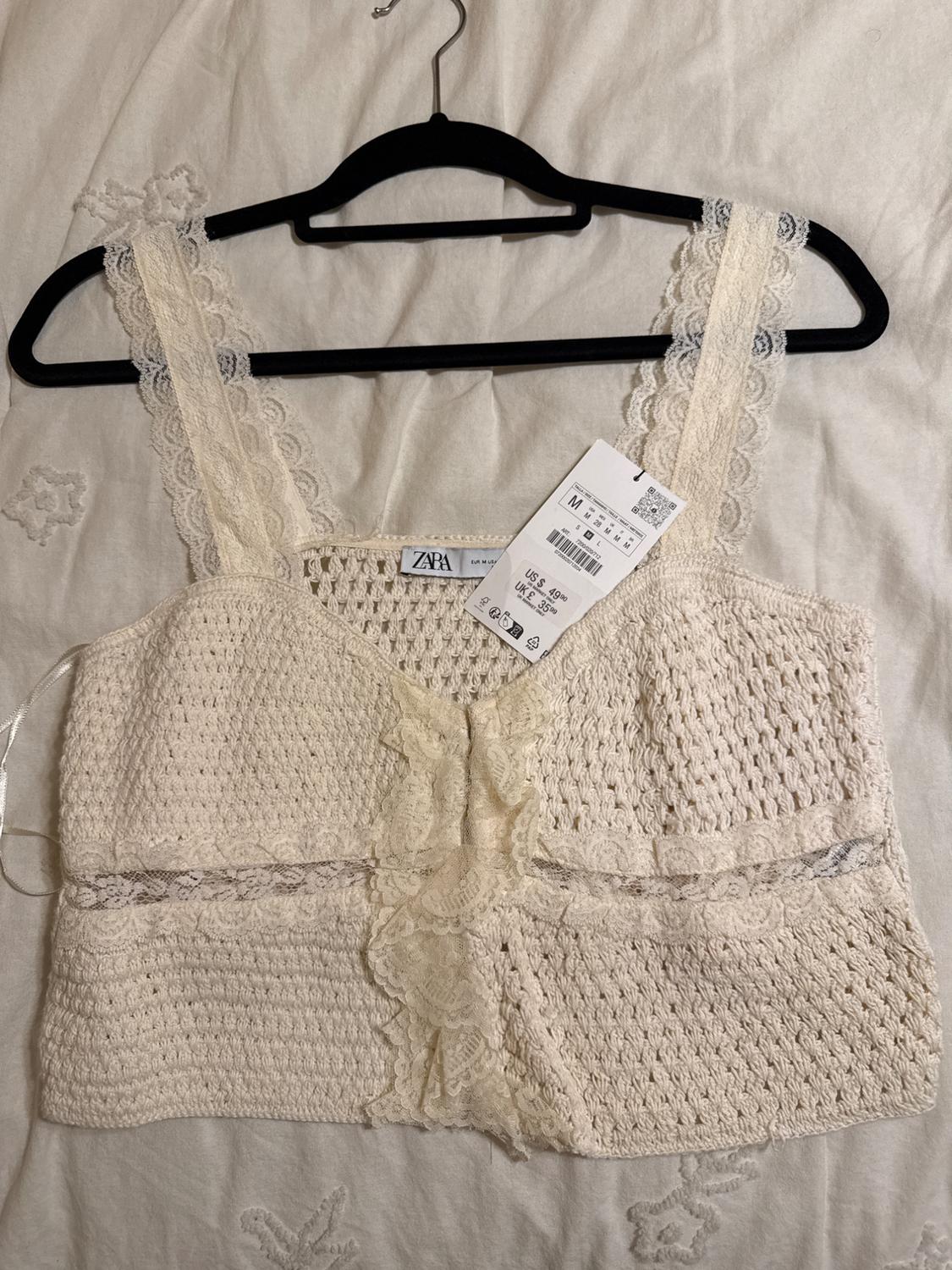White crochet top with lace straps, hanging on a black hanger. Visible label with price and size. White fabric background.