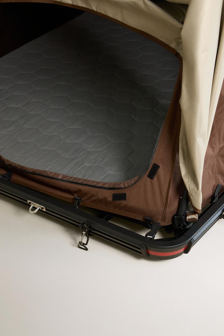 Rooftop tent entrance detail. Shows black base with latches, brown trim with zip, padded grey interior, and beige cover.