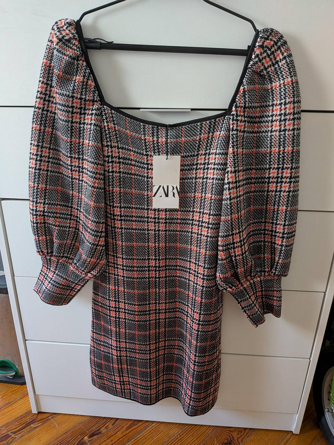 Checked dress with a square neckline and puff sleeves hanging on a hanger in front of white drawers. Label with ZARA text.