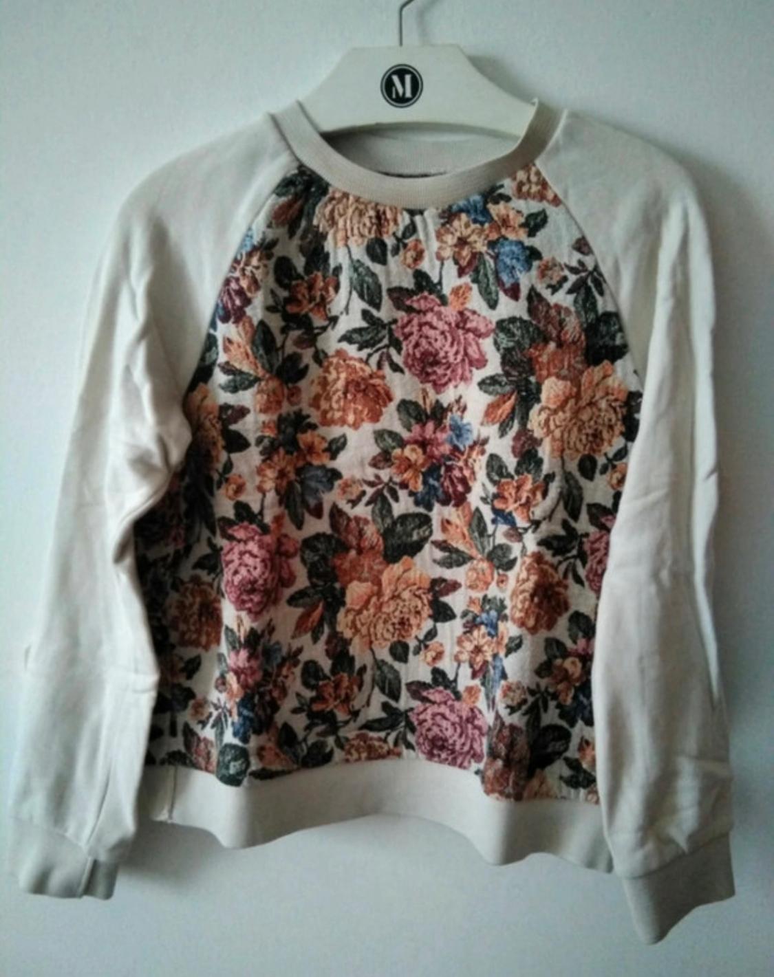 Jumper with long white sleeves and a floral body in shades of pink, orange, and green, hanging on a white hanger with an "M" logo.