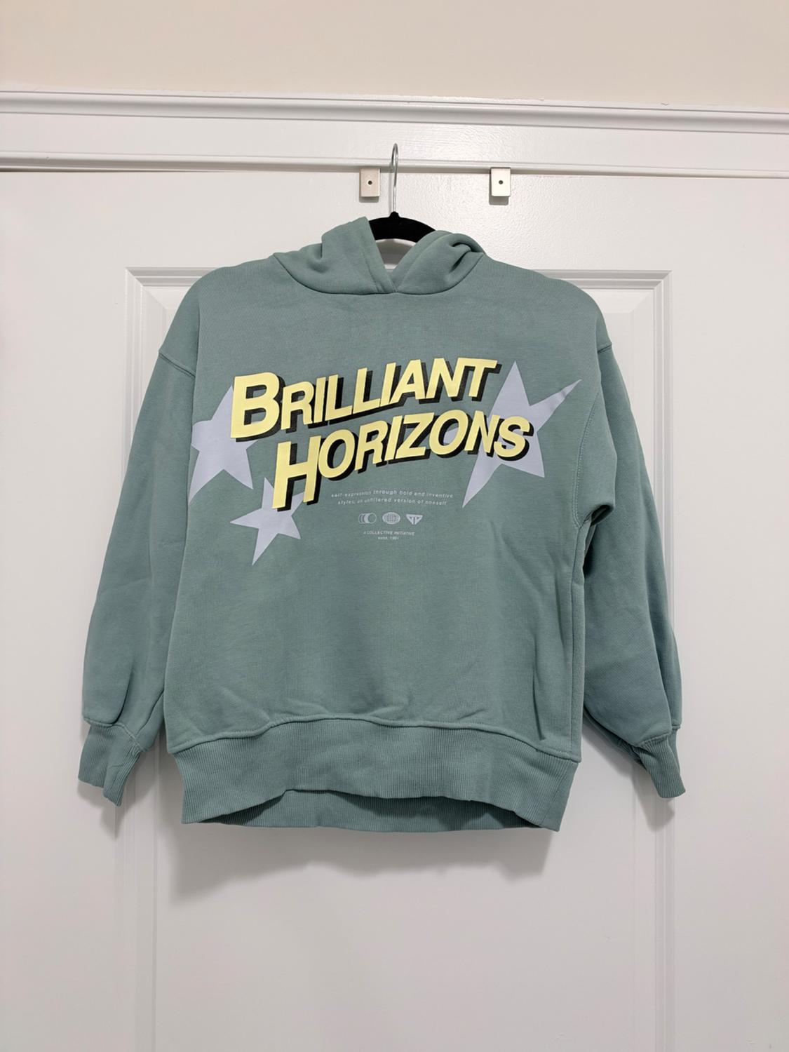 Green hoodie hanging on a white door. In the center, it features gray stars and the yellow text BRILLIANT HORIZONS.