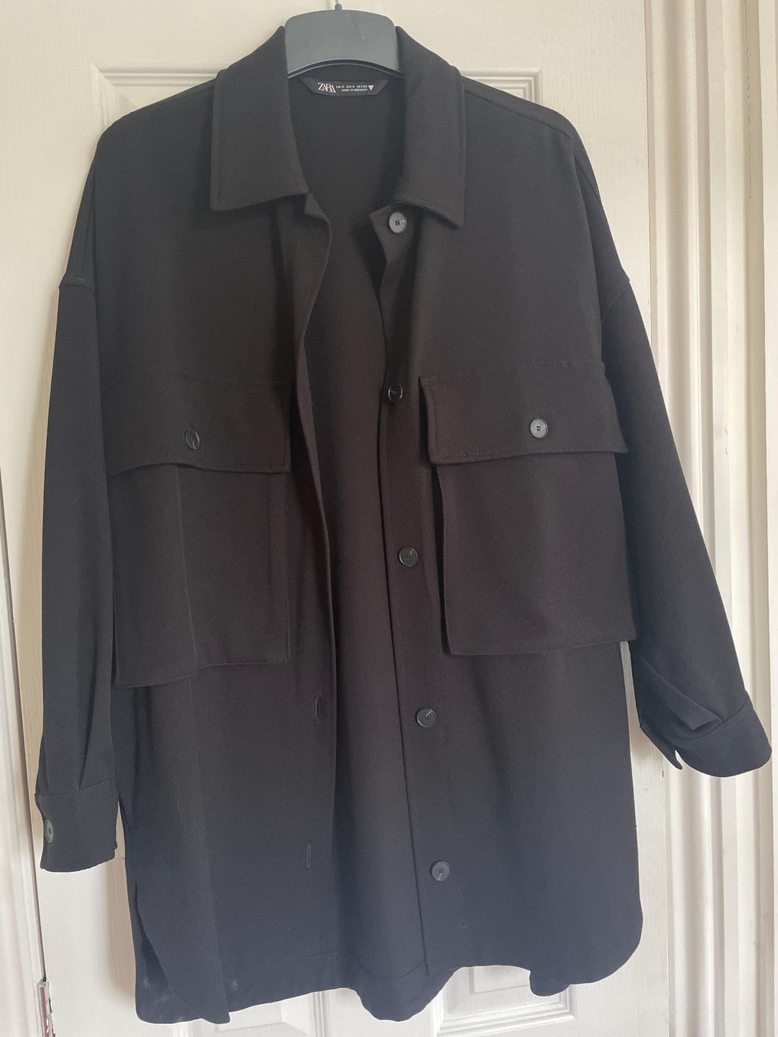 Black coat hanging on a hanger, with large pockets and front buttons. Visible label on the collar. White door background.