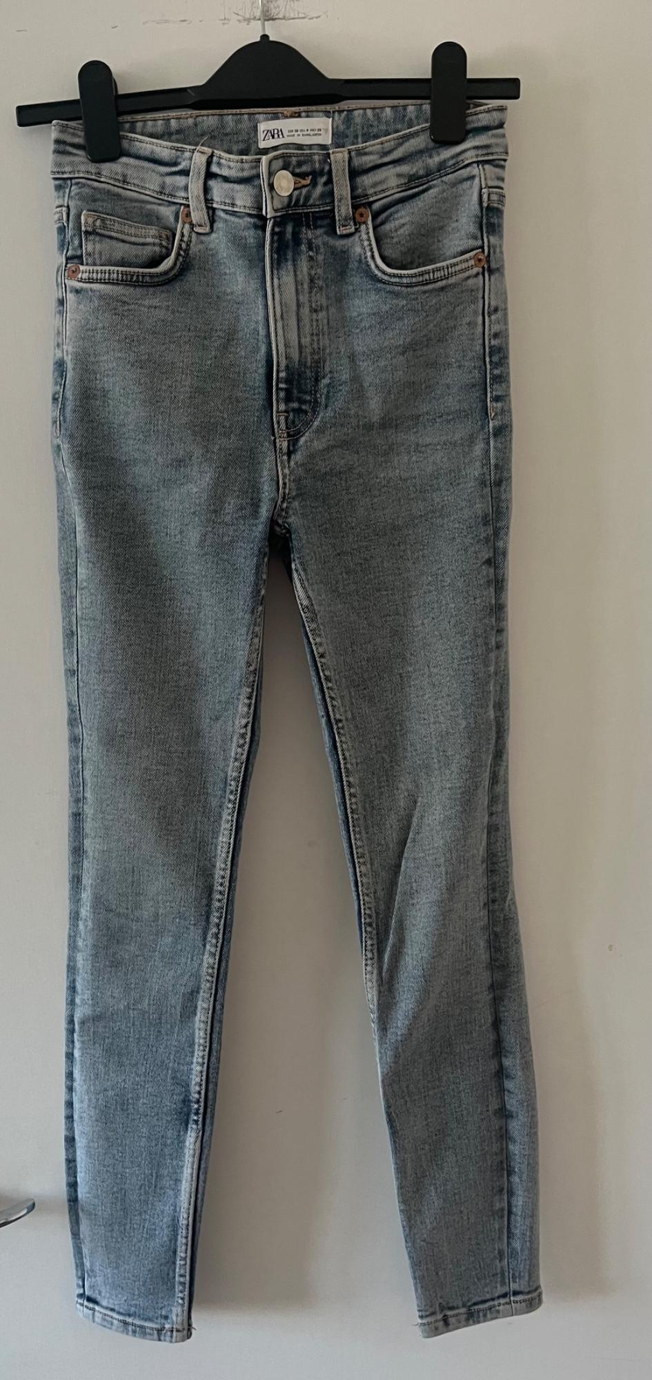 Light blue jeans hanging on a black hanger in front of a white wall. ZARA is written on the label.