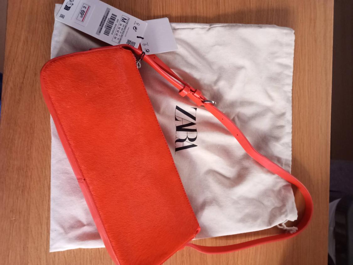 Orange handbag on a white canvas bag with the "ZARA" logo. Price tag visible, showing 69.95. Light wood background.