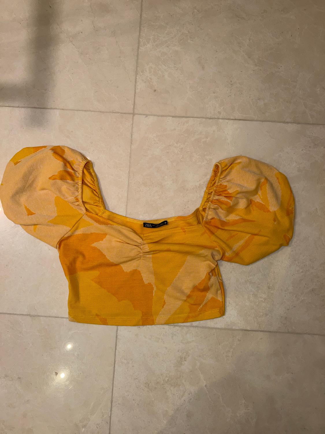 Yellow cropped top with an abstract print and puff sleeves on a light tiled floor. Visible label in the centre.