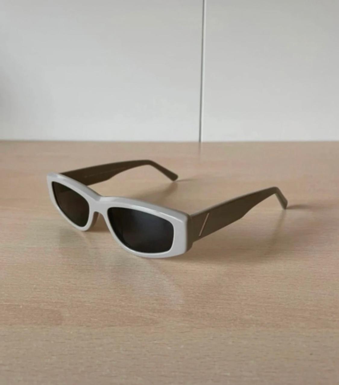 White rectangular-framed sunglasses with black arms on a light wooden surface. Plain white background.
