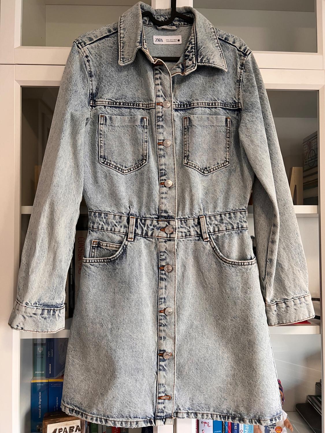 Light blue denim dress with front buttons and chest pockets, hanging in front of a white shelving background.