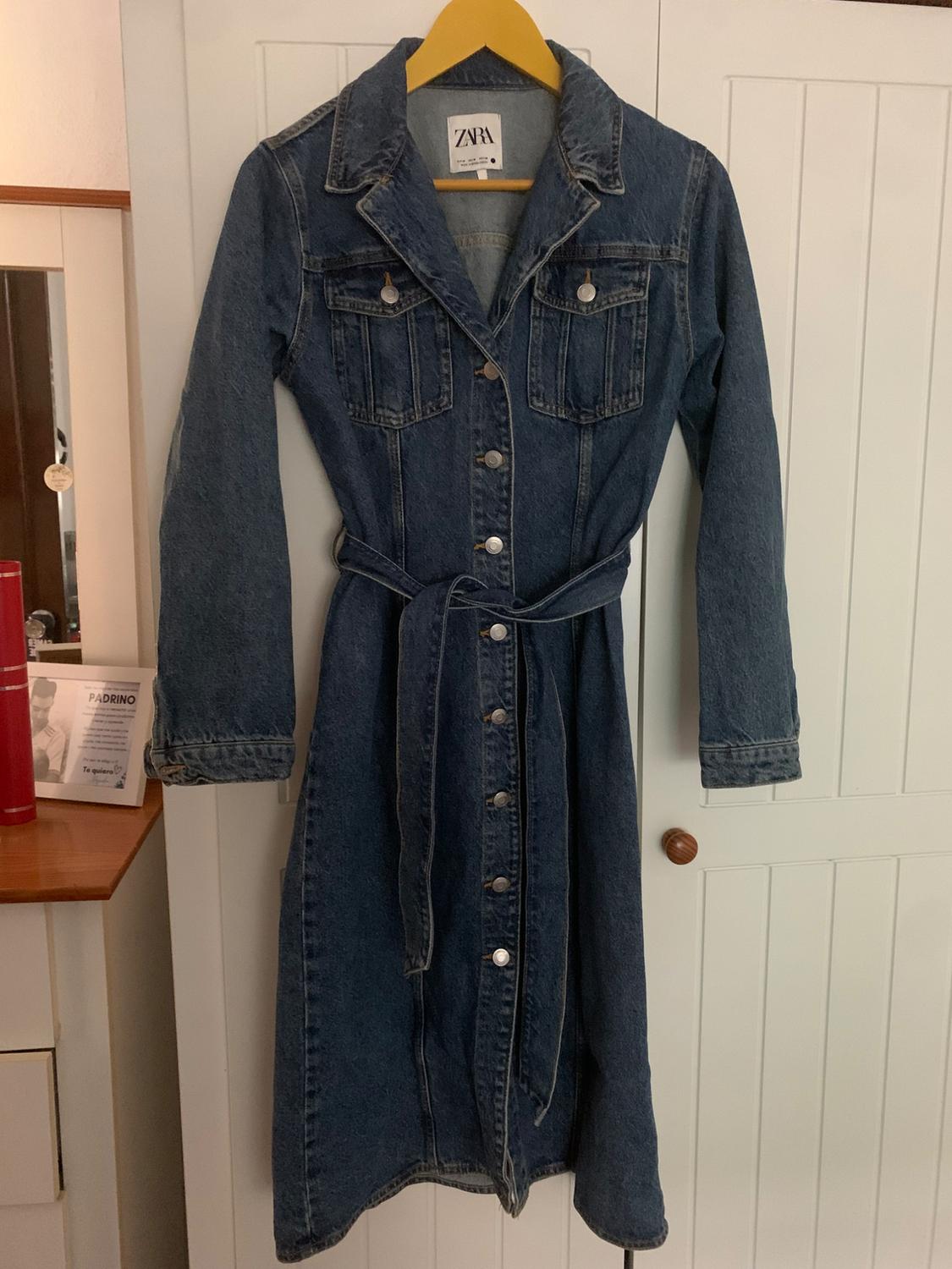 Blue denim dress with buttons and belt, hanging on a yellow hanger. Visible label with text "ZARA". White wardrobe background.