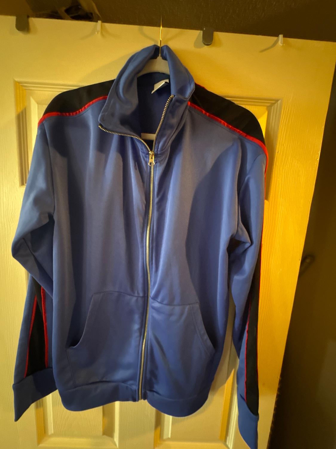 Blue sports jacket with a zip, black and red details on the shoulders, hanging on a light wooden door.