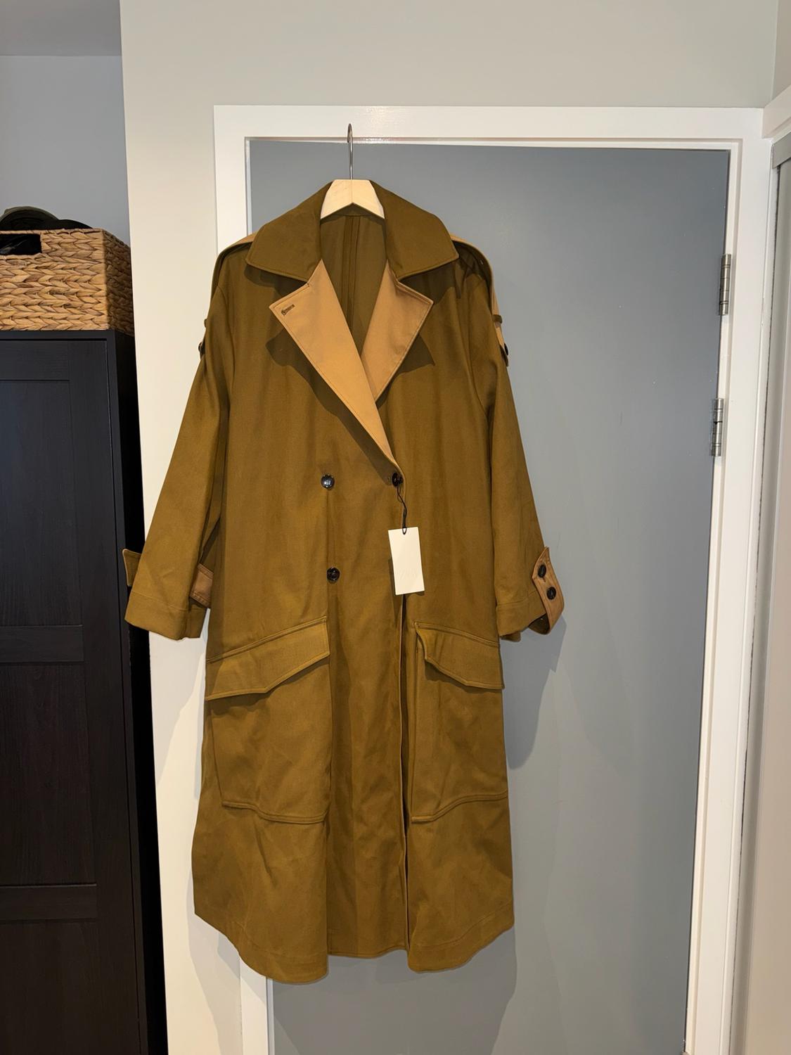Olive green trench coat with beige lapels hanging on a wooden hanger over a grey door. On the left, a piece of furniture with a basket.