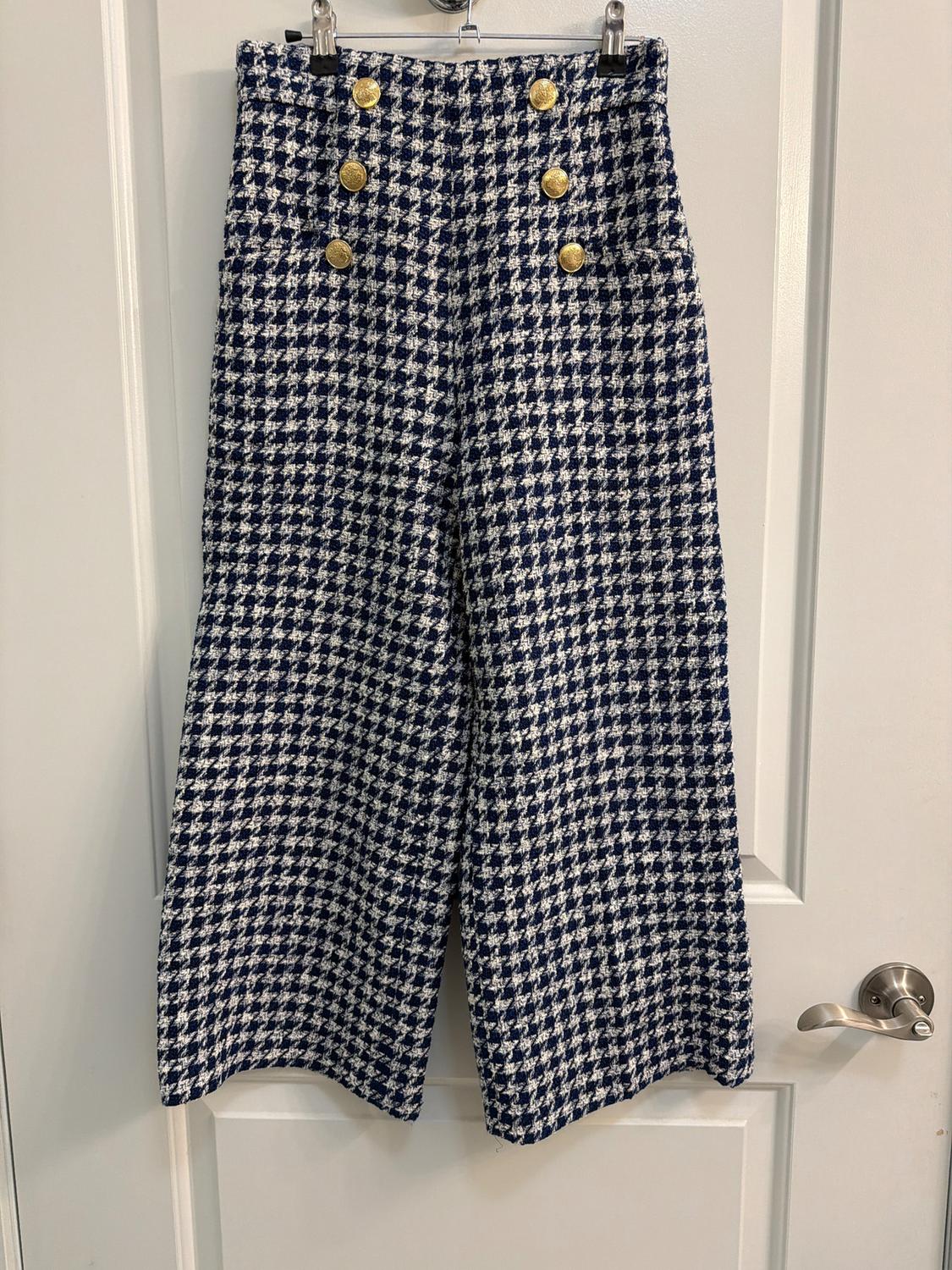Wide-leg pants with a blue and white plaid print, featuring six gold buttons on the front, hanging on a white door.