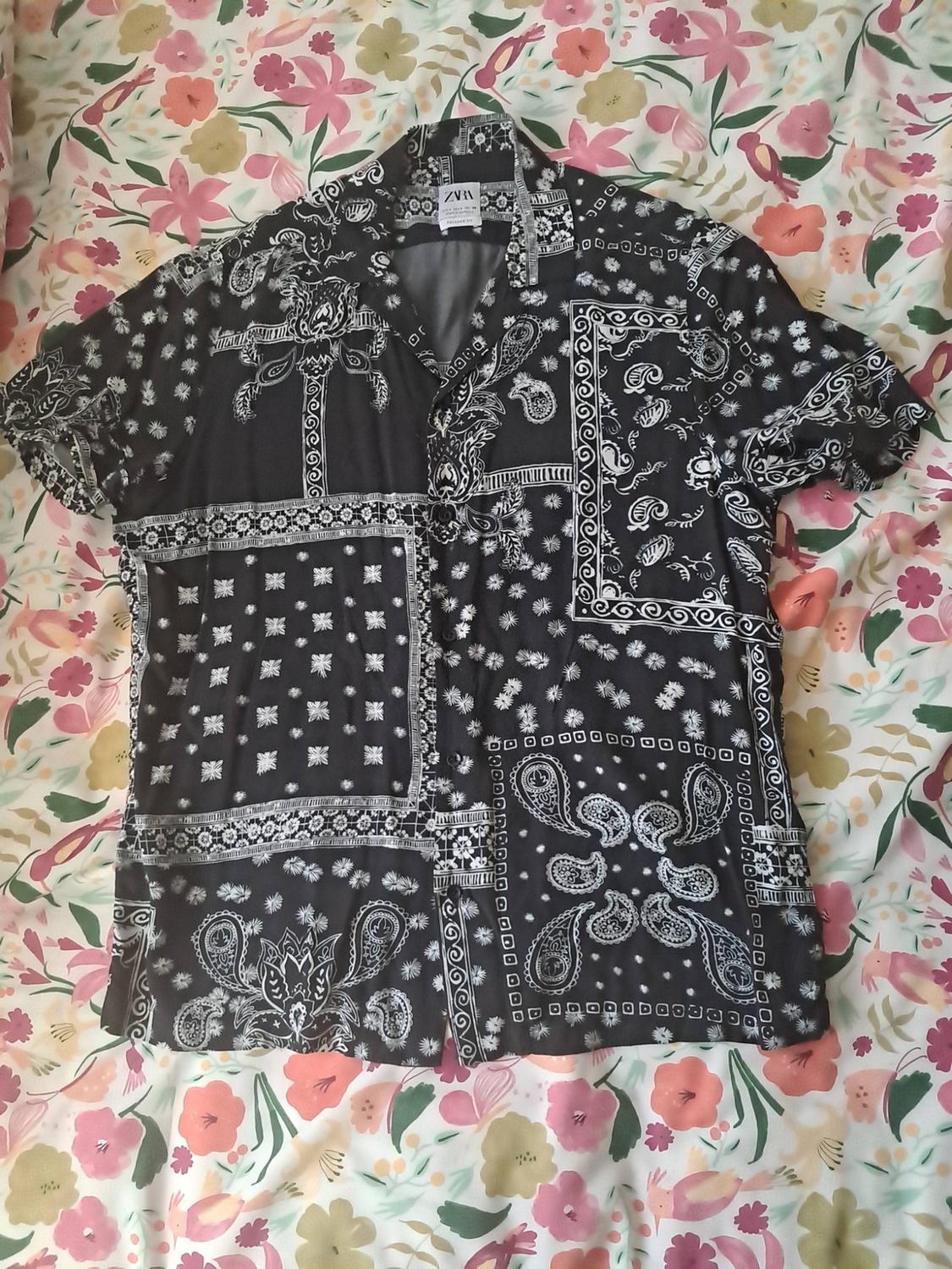 Black short-sleeve shirt with a white bandana print, set against a colourful floral fabric background. ZARA text on the label.