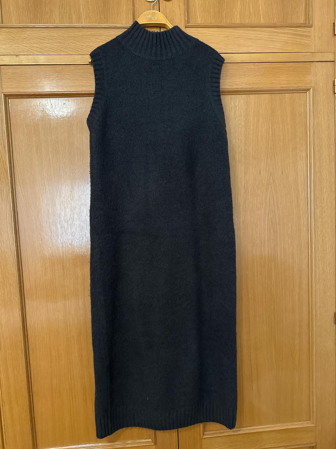 Black, sleeveless, chunky knit dress with a high neck, hanging on a wooden hanger in front of a light wood wardrobe door.