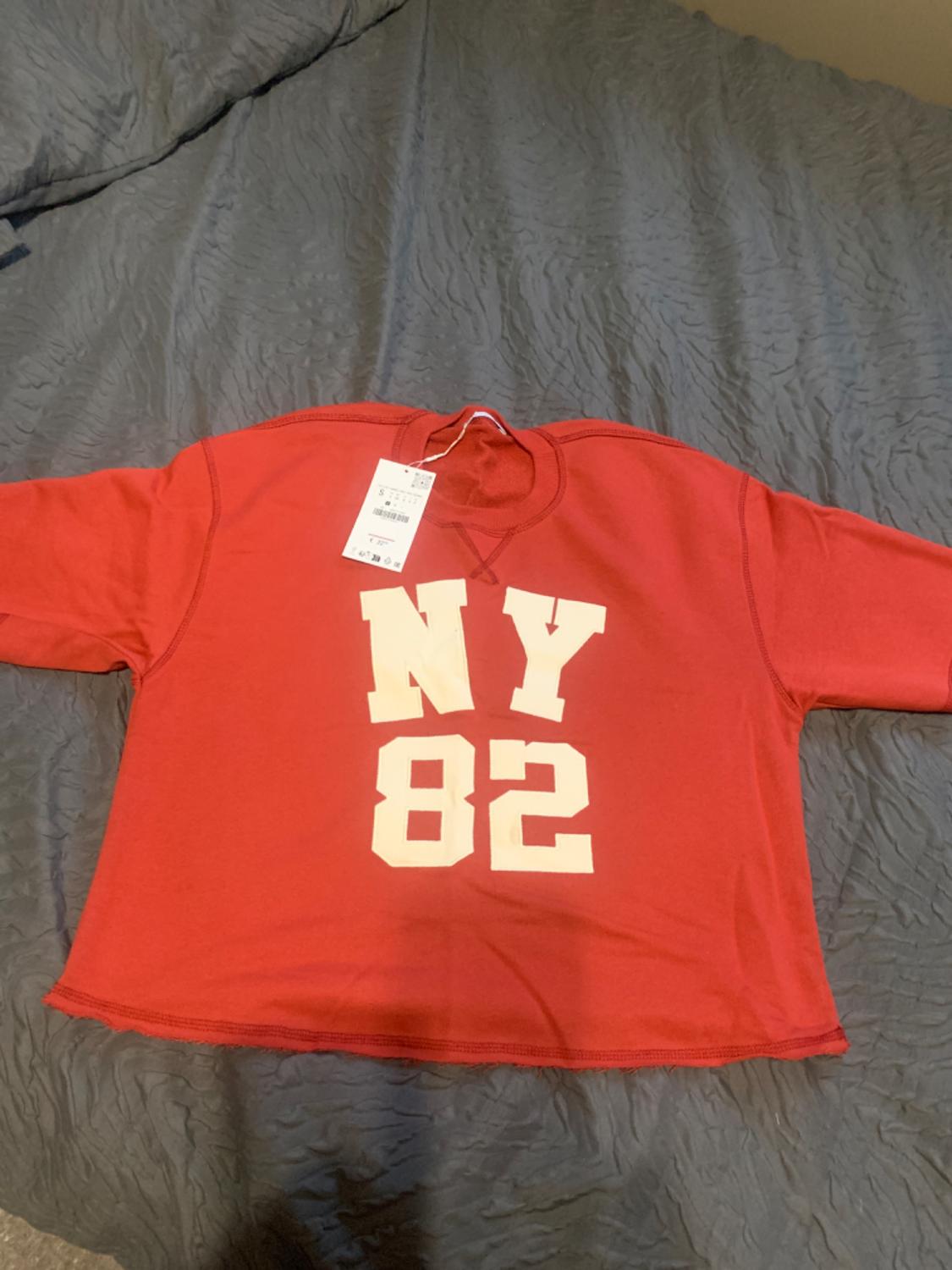 Red T-shirt with "NY 82" text in white, on a bed with a grey quilt. Label hanging from the neck.