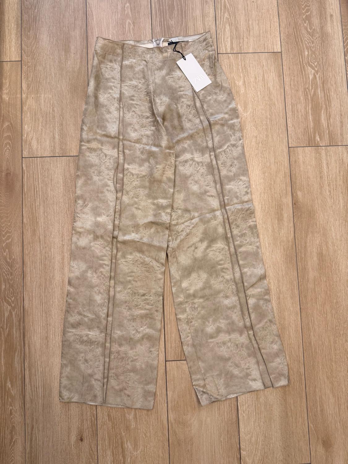 Beige trousers with a crinkled texture and front seams, laid out on a wooden floor. Label with the text ZARA.