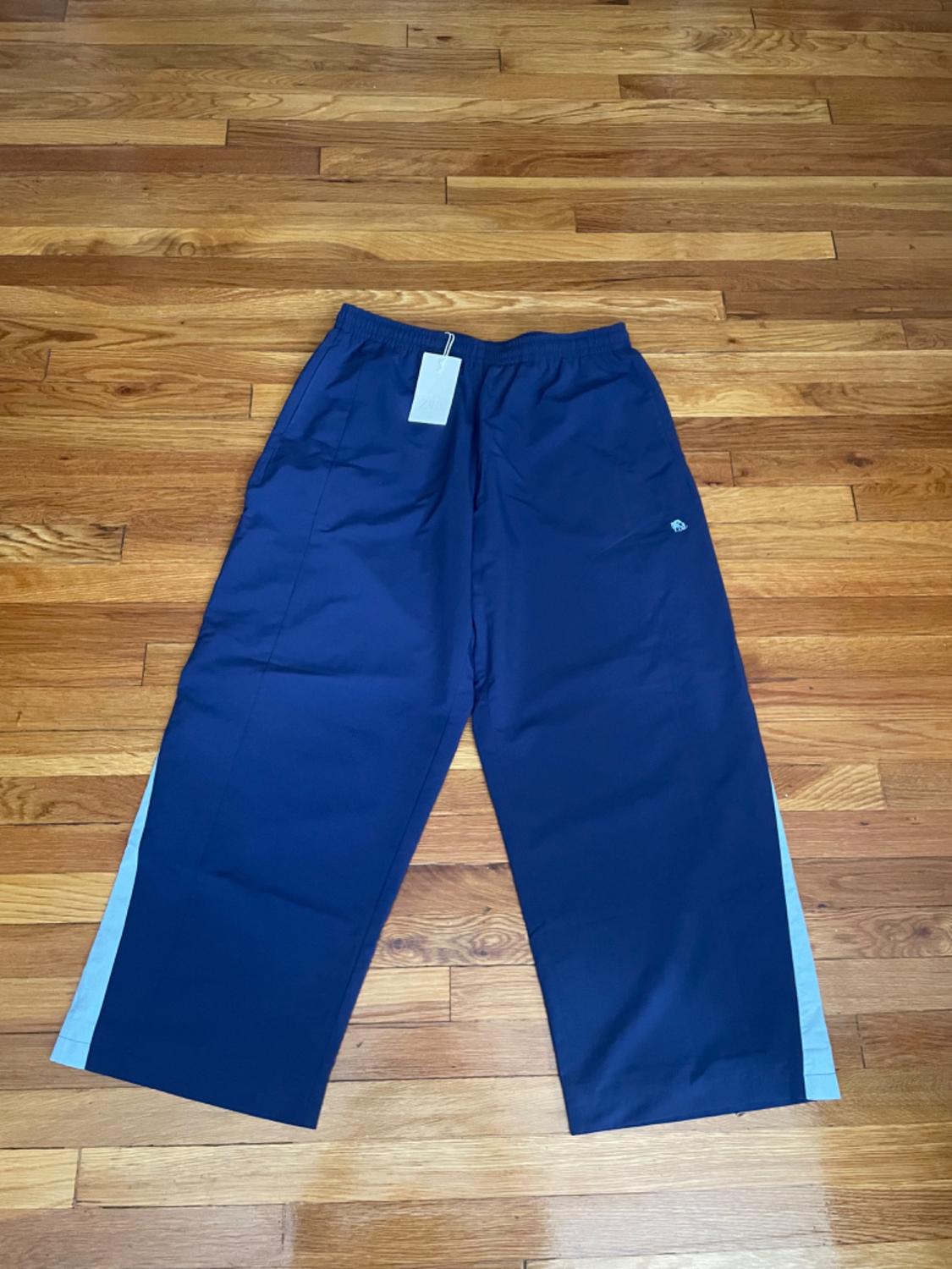 Wide navy blue tracksuit bottoms with light blue side stripes on a wooden floor. They have an elasticated waistband and a white label.