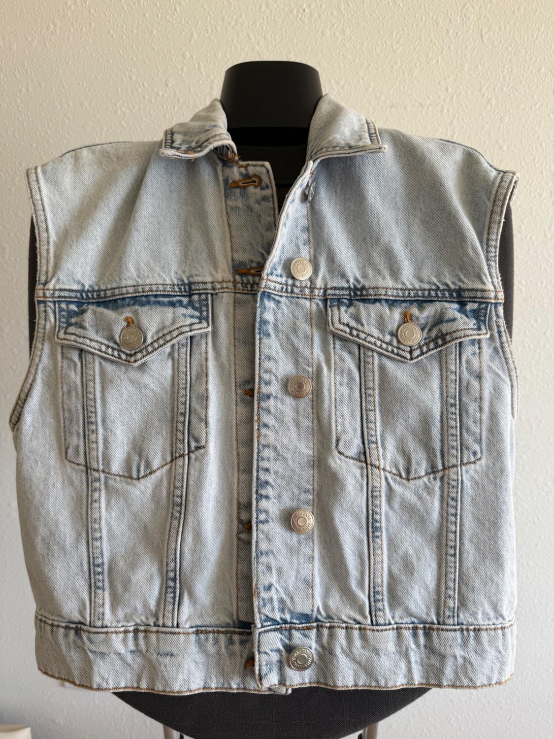 Light blue sleeveless denim waistcoat on a black mannequin, with metal buttons and front flap pockets.