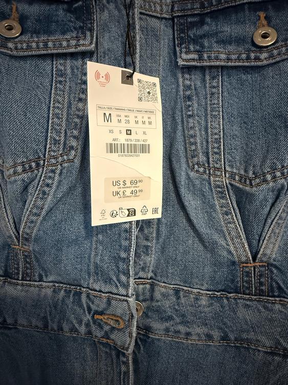 Price tag hanging from a blue denim jacket. Visible text: "US$ 69.90, UK£ 49.90". Metal buttons and visible stitching.