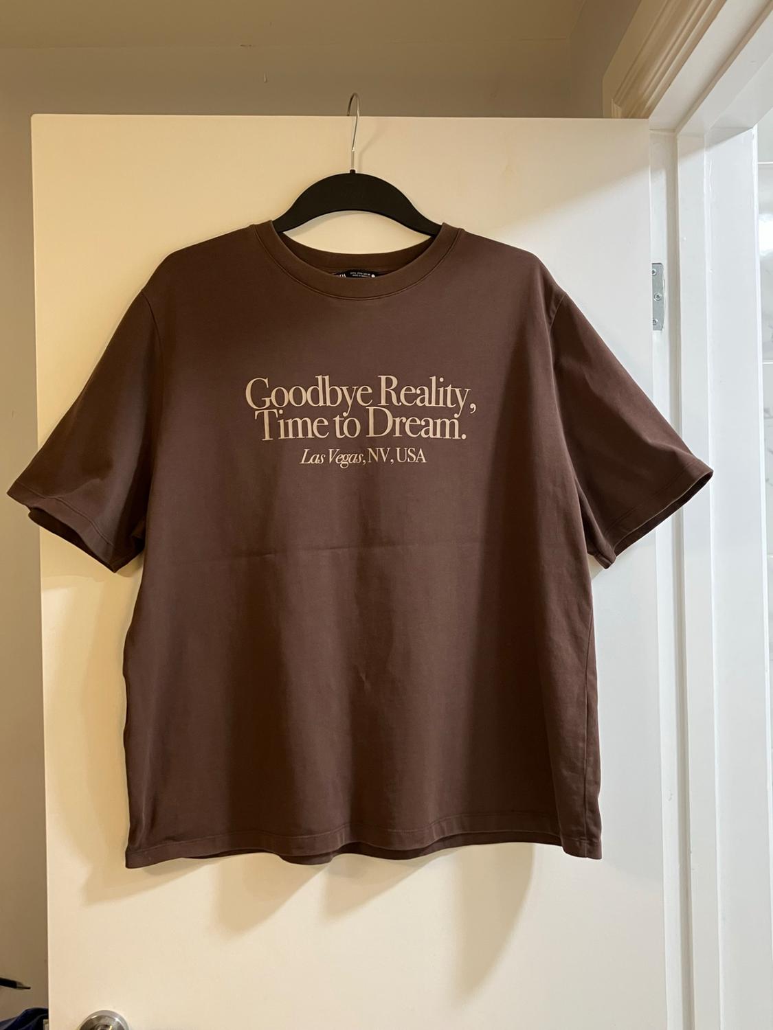 Brown short-sleeved T-shirt hanging on a white door with the text: 'Goodbye Reality, Time to Dream. Las Vegas, NV, USA'.