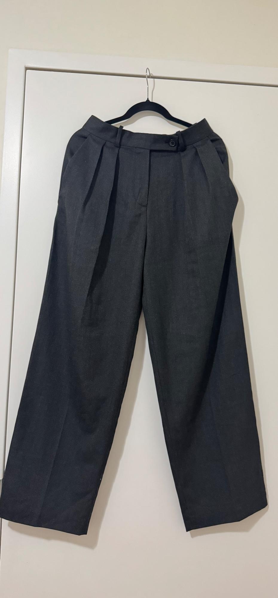 Gray wide-leg pants hanging on a black hanger over a white door. Design with pleats and side pockets.