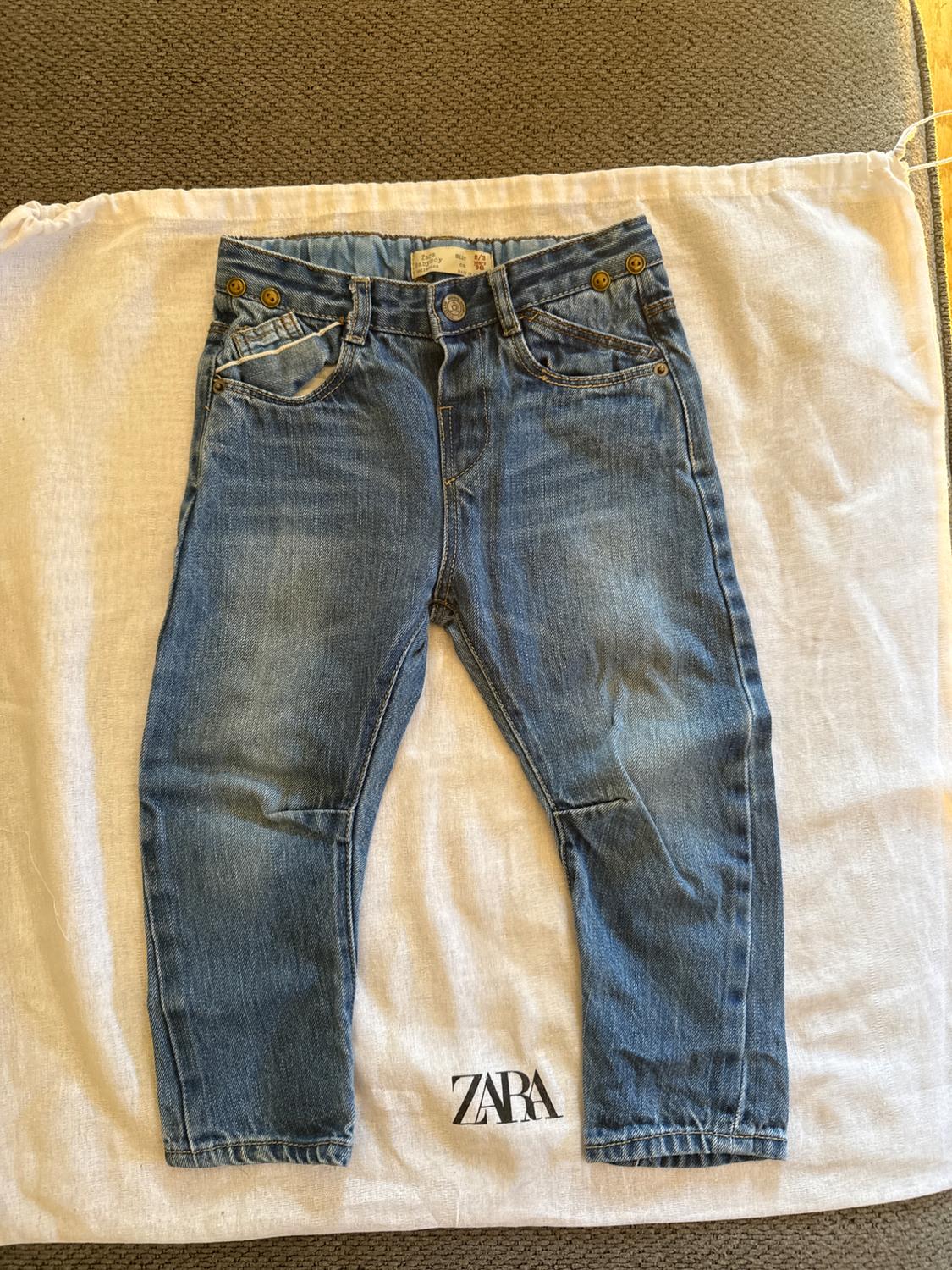 Blue children's jeans on a white bag with the Zara logo, placed on a grey fabric surface.