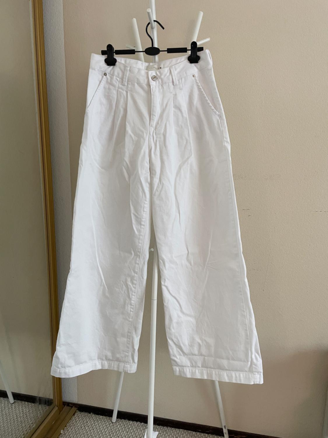 White wide-leg pants hanging on a white hanger, with a beige wall background and a mirror on the left.