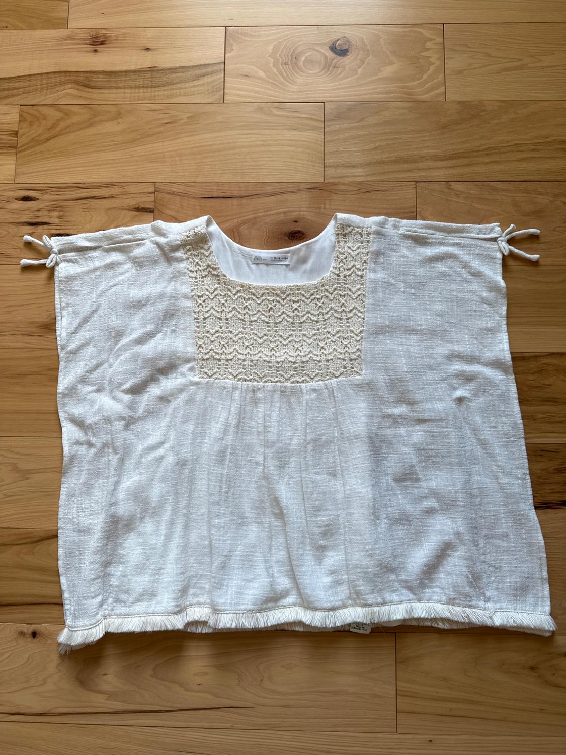 White blouse made of light fabric with lace details on the chest, ties on the sleeves, and fringe on the bottom hem, over light wood flooring.