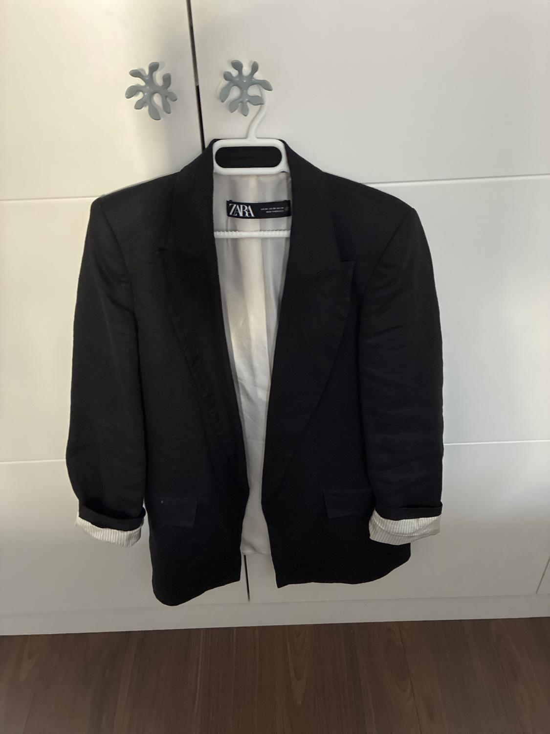 Open black blazer hanging in a white wardrobe. Sleeves rolled up with a striped lining. The label inside reads Zara.