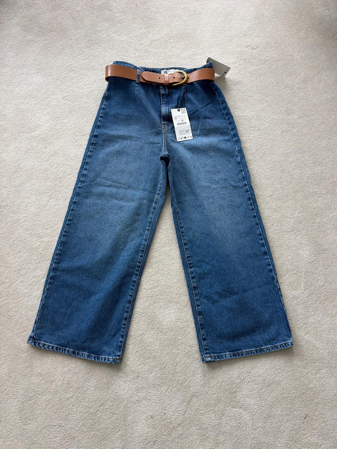 Blue wide-leg jeans with a brown belt and tags, laid out on a light-coloured rug.