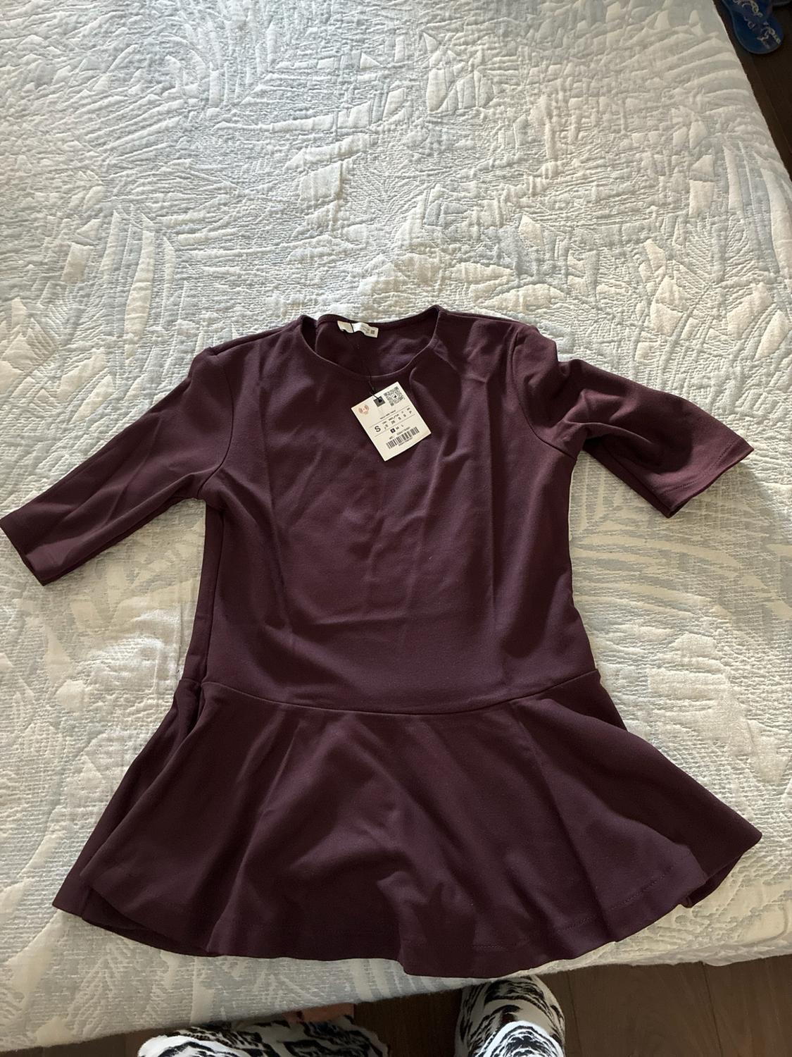 Burgundy short-sleeved top with a frilled hem and tag, on a bed with a light-coloured textured bedspread.