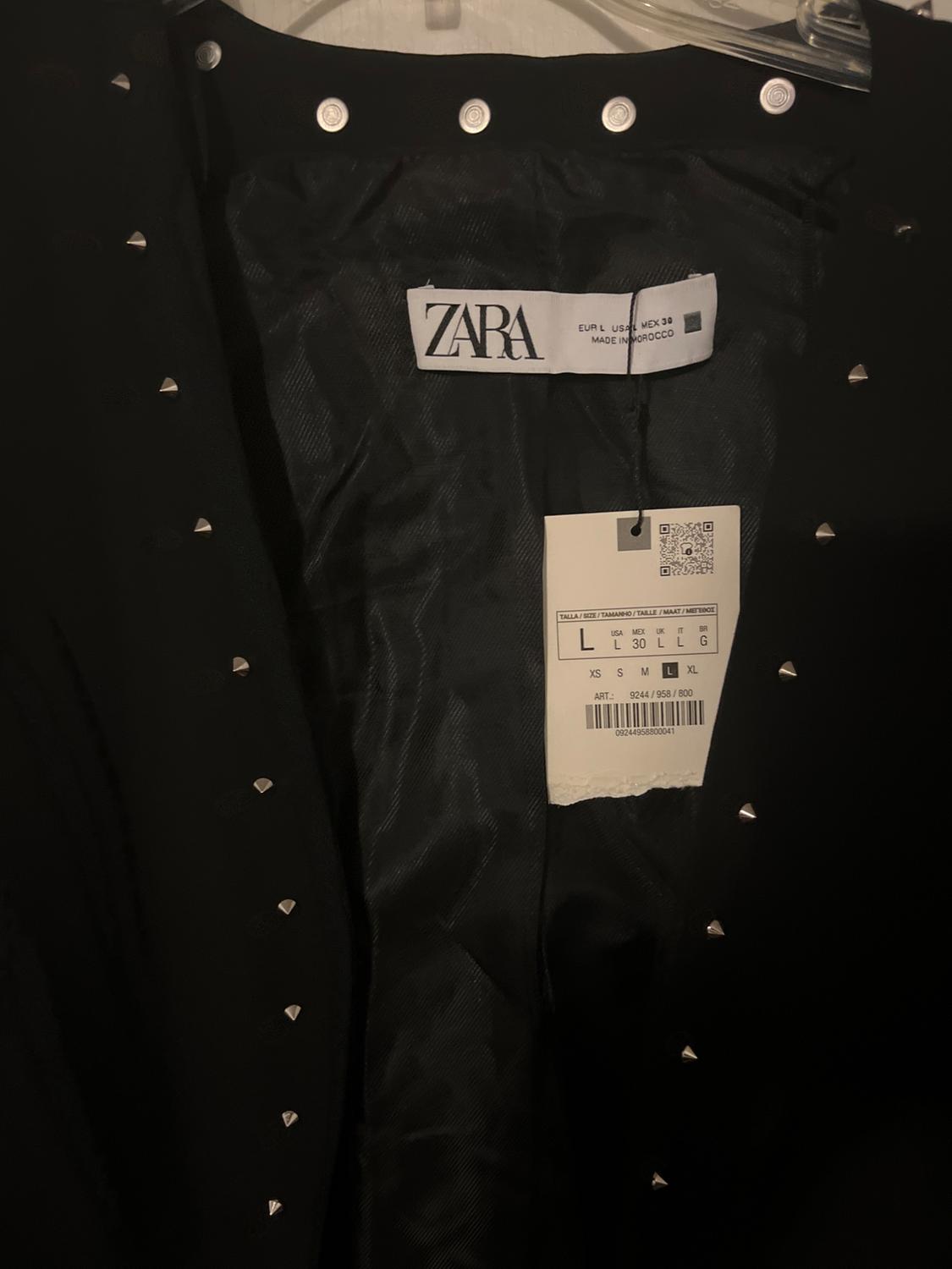 Inner label of black jacket with studs and visible Zara label. Text: "ZARA EUR L USA L MEX 30 MADE IN MOROCCO".