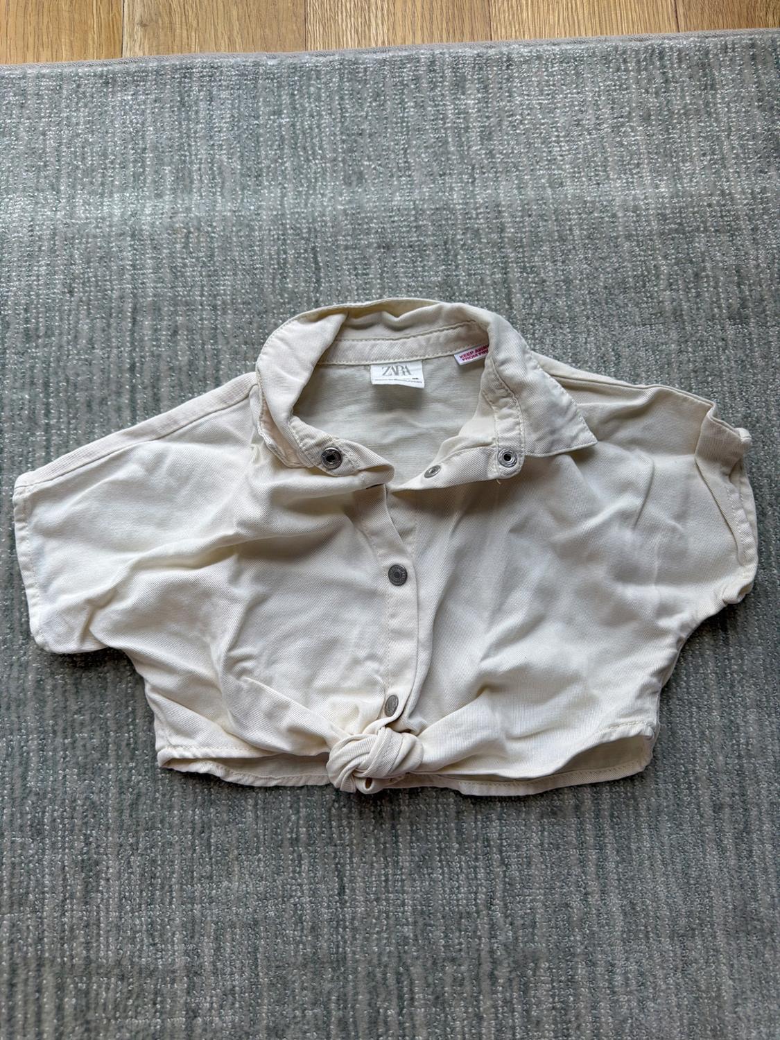 Short beige shirt with buttons and a front knot, on a grey rug. Visible label on the collar. Light wood background.