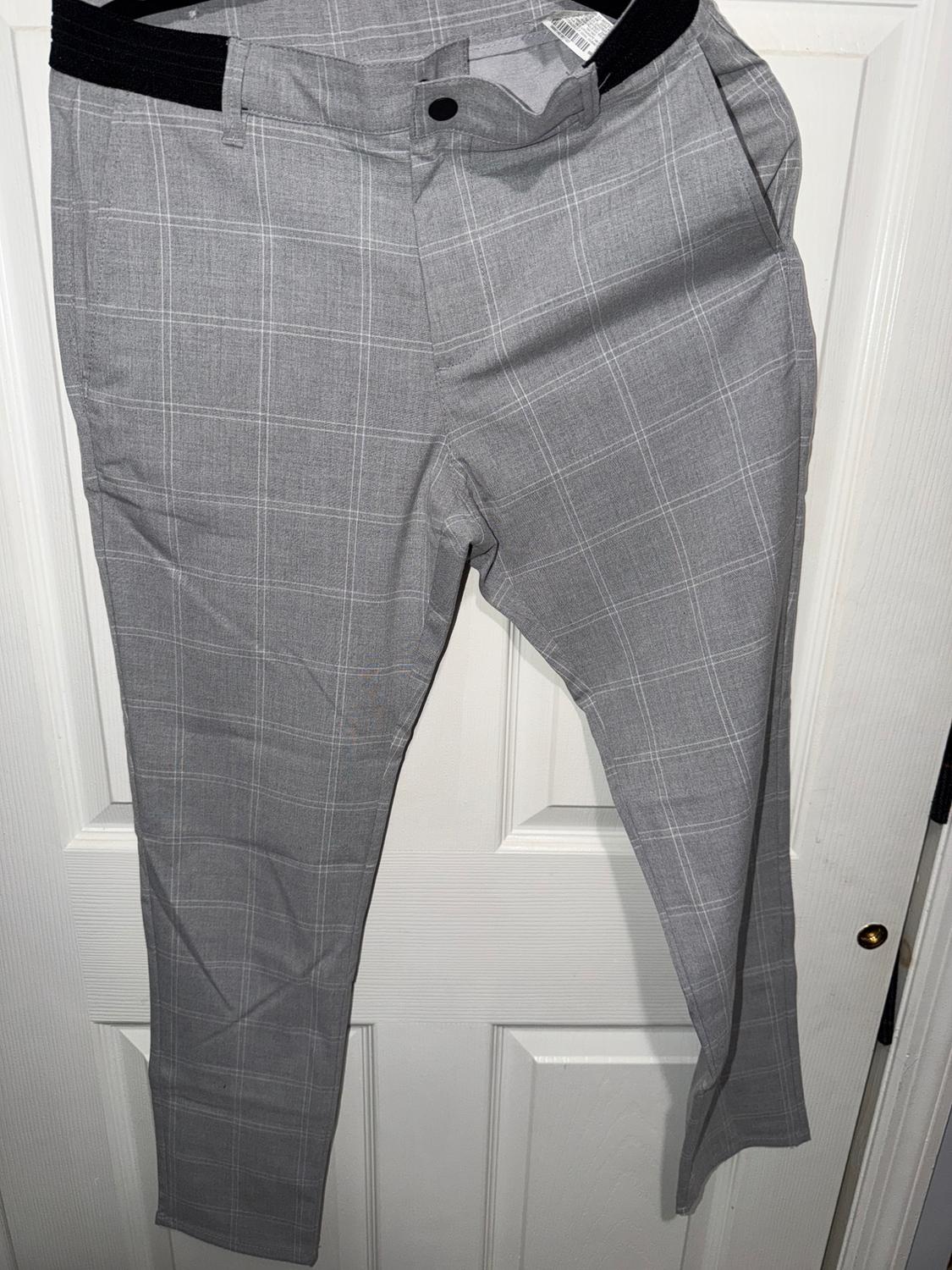 Grey trousers with a checked pattern, hanging on a white door. Waistband with a black button and visible side pockets.