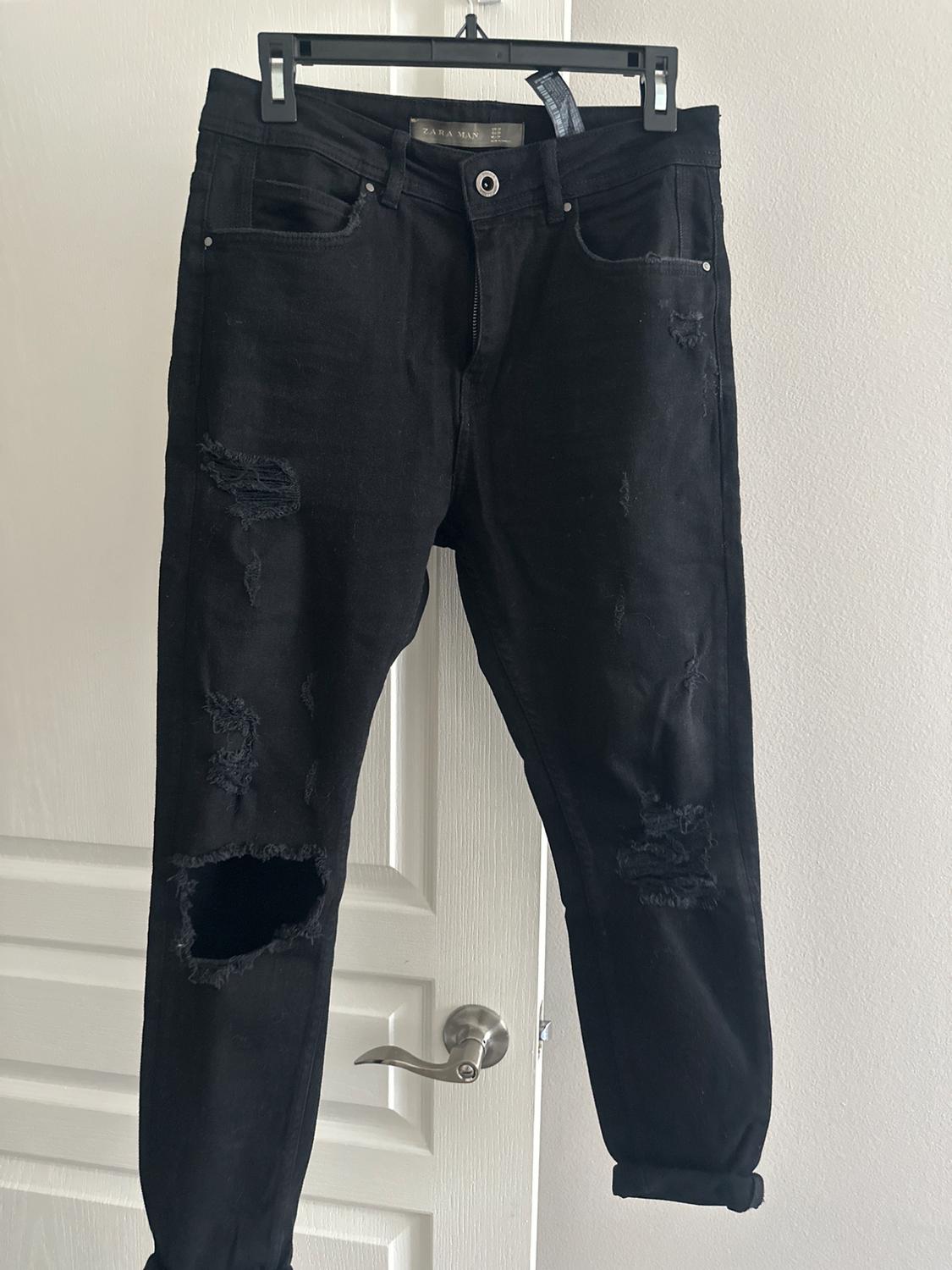 Worn black trousers hanging on a hanger in front of a white door. Visible tag: "ZARA MAN".