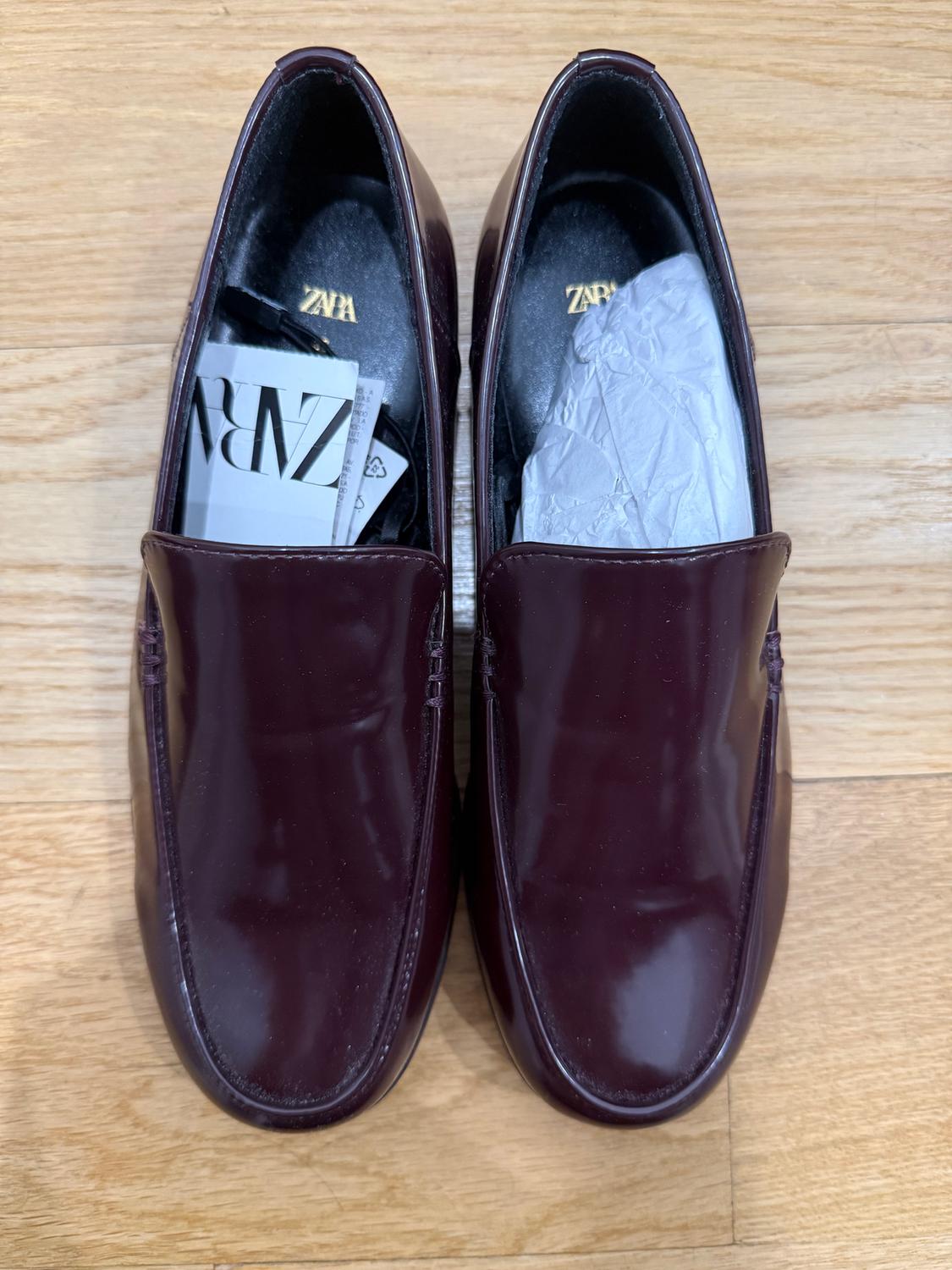 Burgundy patent leather shoes on a wooden floor, with paper and cards inside. Visible text: "ZARA".