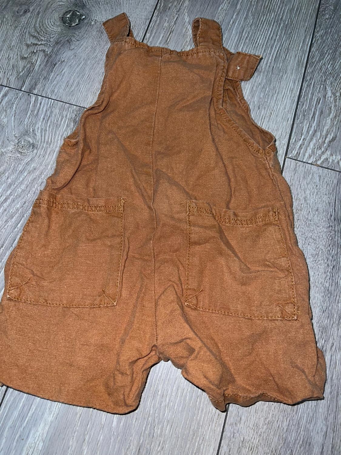 Brown fabric dungarees with straps and two front pockets, placed on a grey wooden floor.