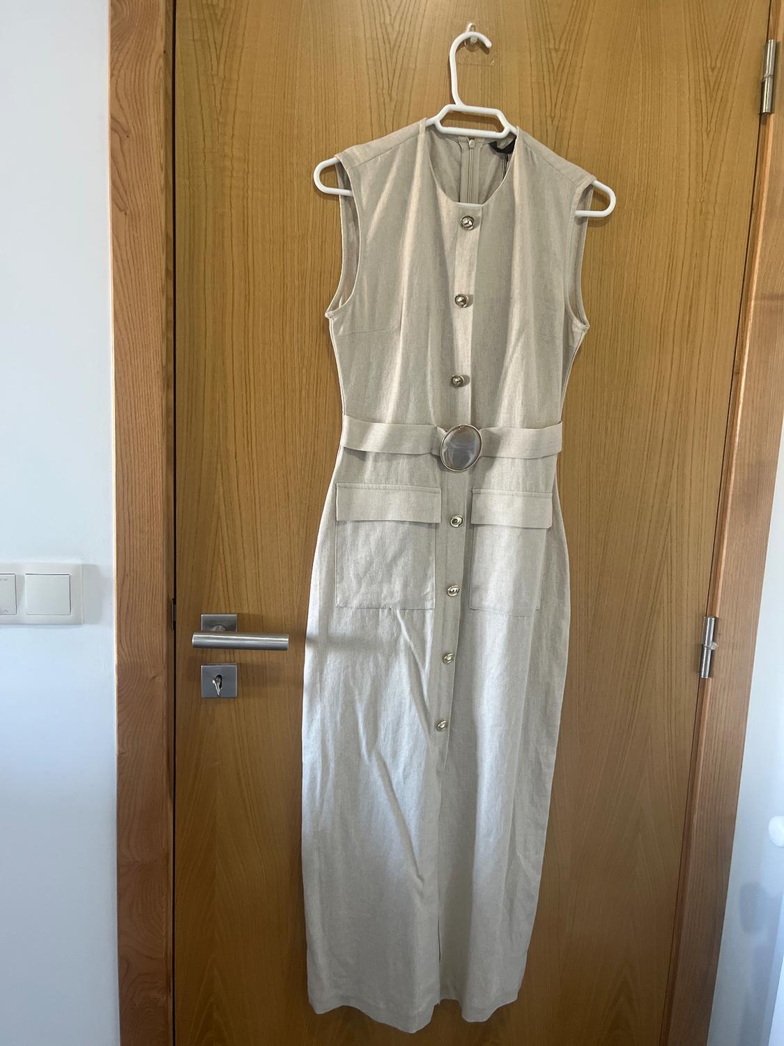 Beige sleeveless dress with front buttons and a round buckle belt, hanging on a light wooden door.
