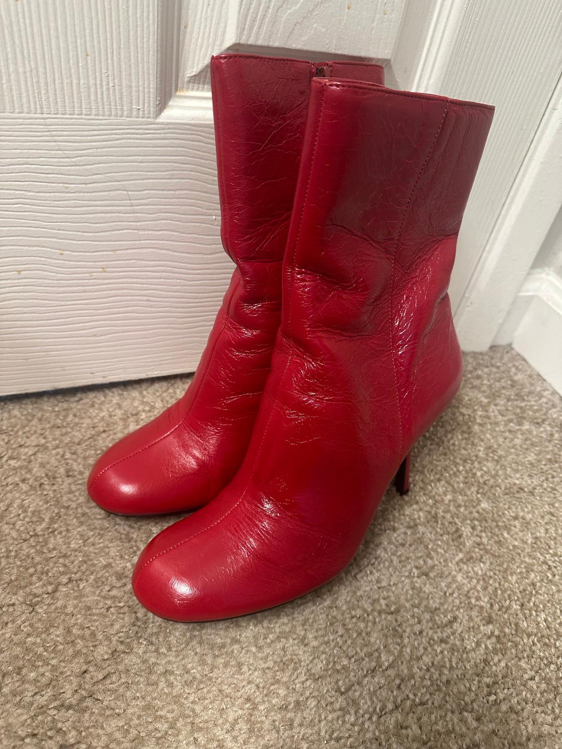 Red, mid-heel ankle boots on a beige rug, next to a white door. The material has a glossy finish.