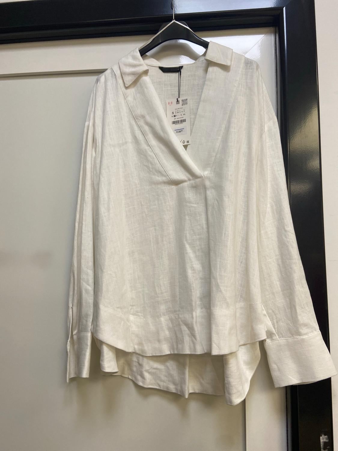White long-sleeved shirt with a V-neck and an asymmetric hem, hanging on a black hanger on a white door with a black frame.