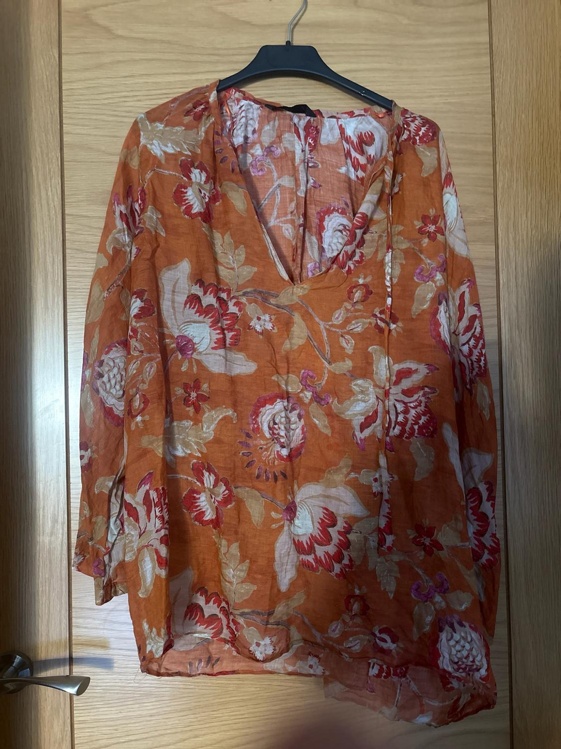 Orange long-sleeved V-neck blouse with a white and red floral print, hanging on a hanger on a wooden door.