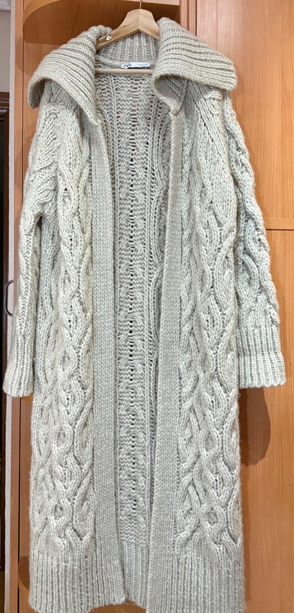 Long, chunky knit beige coat with a braided design, hanging on a wooden hanger against a wooden wardrobe background.