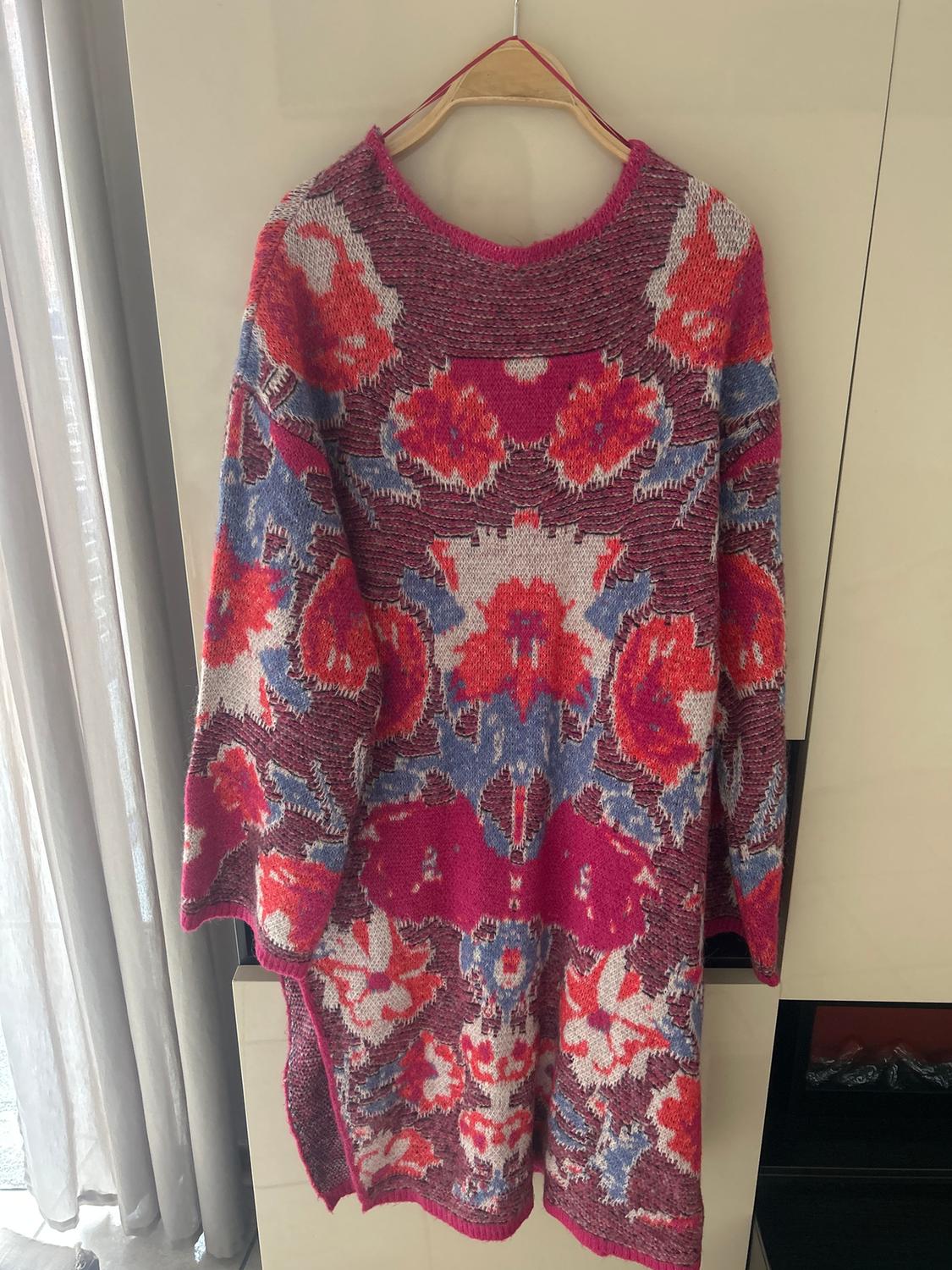 Jumper hanging on a hanger, with a floral design in red, blue, and white tones, on a pink and grey background. Curtains on the left.